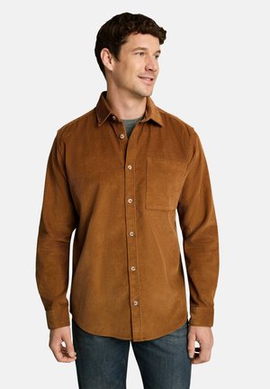 Mann trägt ein braunes Cordhemd über einem grauen T-Shirt und dunkelblauen Jeans, schaut mit einem leichten Lächeln zur Seite.