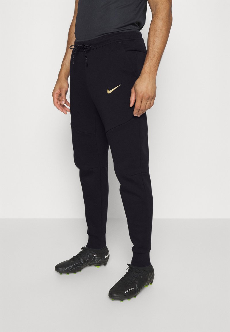 Jogging noirs athlétiques avec une coupe slim, ornés d'un logo Nike doré, d'une taille à cordon de serrage et de poches latérales. Portés avec des chaussures à crampons noires.