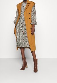 Senapsgul ärmlös trenchcoat med två svarta knappar, lagd över en blommig klänning i grönt, brunt och kräm. Bruna högklackade ankelskotor.