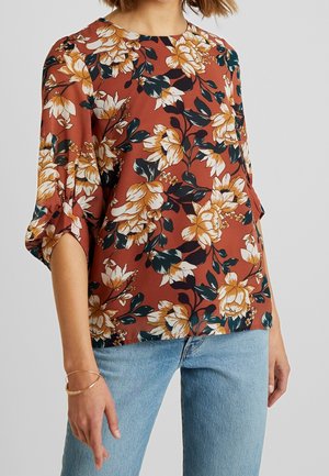 Blusa floreale in ruggine con grandi fiori crema e verdi. Vestibilità ampia, con maniche a tre quarti e spacchi laterali. Tessuto leggero.