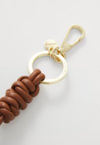Portachiavi in pelle marrone con texture annodata, dotato di un anello circolare in tonalità oro e di un gancio in metallo per il fissaggio.