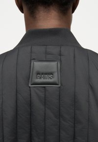 Πίσω όψη ατόμου που φοράει μαύρο μπουφάν με καπιτονέ και ριμπ λεπτομέρεια στον γιακά, με μαύρο ετικέτα "RAINS" στο πάνω μέρος της πλάτης.