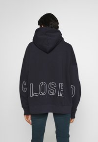 Marinblå oversize-hoodie med en stor vit "CLOSED"-grafik på baksidan, utrustad med dragsko i huvan och ribbad nederkant. Tillverkad i bomullsblandning.