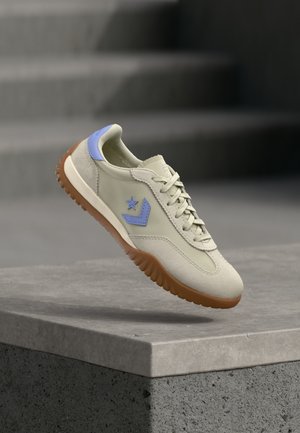Beżowy sneaker z niebieską gwiazdą i logo w kształcie szewronu, gumowa podeszwa i sznurówki, pokazany w powietrzu nad betonową krawędzią z rozmytymi szarymi schodami w tle.