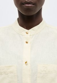 Közelkép egy személyről, aki krémszínű, gombos ingben van, amelynek mandzsettás gallérja és két mellzsebje van, az egyik rókalogóval hímzett.