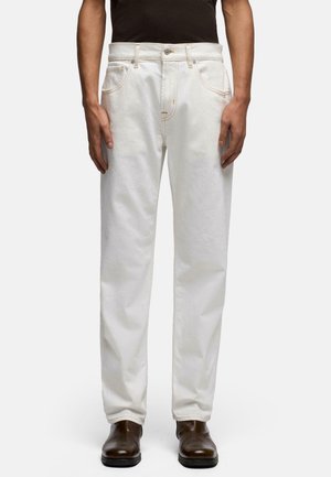 Mann trägt weiße Jeans mit geradem Bein, schwarzes Hemd und braune Lederschuhe, steht vor einem schlichten Hintergrund.