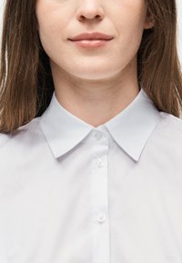 Κάτω μέρος προσώπου και λαιμός γυναίκας με ανοιχτόχρωμο δέρμα, φοράει λευκό πουκάμισο με κουμπιά και γιακά, με μακριά καστανά μαλλιά που φαίνονται στα πλάγια.