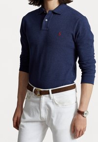 Tmavomodré polo tričko s dlouhým rukávem a límečkem, se malým červeným logem, spárované s bílými kalhotami a hnědým koženým opaskem.