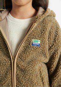 Persona che indossa una giacca in fleece beige con una texture, con la cerniera parzialmente aperta, che mostra una maglietta bianca a girocollo sottostante.