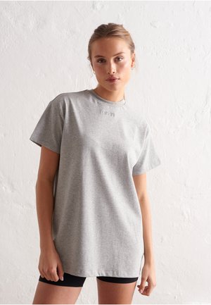 Jeune femme portant un t-shirt gris à manches courtes et ample, et un short noir, debout contre un mur blanc texturé.