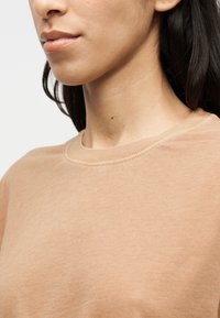 Sweat-shirt en coton marron clair avec un col rond et une texture lisse, présentant des détails discrets de coutures le long de l'encolure.