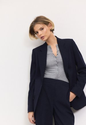 Jeune personne aux cheveux blonds courts portant un blazer bleu marine, une chemise grise boutonnée et un pantalon bleu marine assorti, debout devant un fond blanc.