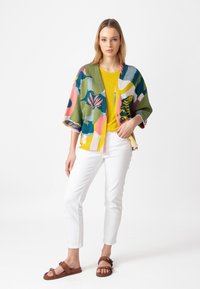 Kleurige, gebreide cardigan met patroon en korte mouwen, gelaagd over een felgeel topje. Witte slim-fit broek en bruine sandalen maken de look compleet.