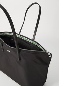 Black tote bag made of nylon, featuring two long handles, zip closure, and a green logo lining with a small embroidered logo on the front.