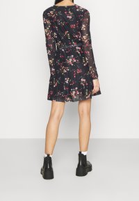Schwarz-blumiges Kleid mit langen transparenten Ärmeln und taillierter Taille. Verziert mit kleinen bunten Blumen und einem fließenden Rockdetail. Schwarze Knöchelstiefel.