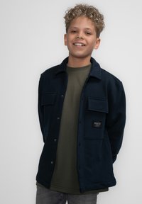 Donkerblauw flanellen jasje met knoopsluiting, twee borstzakken en een merklabel. Draag het over een groene t-shirt. Effen achtergrond.