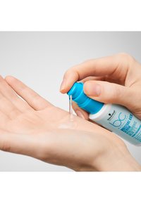 Un gel clair est distribué d'une bouteille à bouchon bleu dans une paume ouverte. La bouteille a un corps blanc avec du texte et des accents bleus.