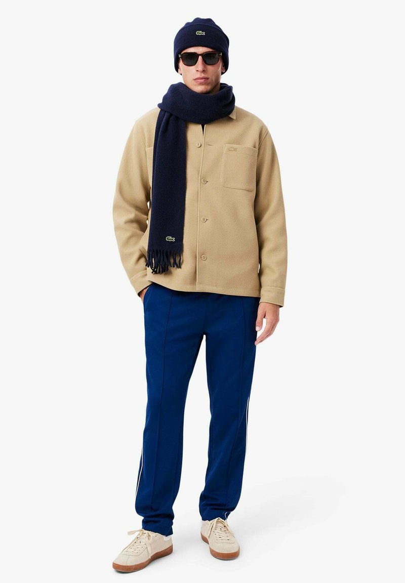 Veste en polaire beige, écharpe bleu marine avec des franges, pantalon bleu marine et baskets beiges. Les accessoires comprennent un bonnet bleu marine et des lunettes de soleil sombres.