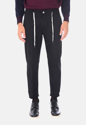 Pantaloni neri a sigaretta con vita elasticizzata, cordino bianco e tasche laterali, abbinati a scarpe nere e un maglione scuro.