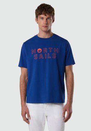 Jeune homme aux cheveux courts et bouclés portant un t-shirt bleu avec le texte rouge "NORTH SAILS" et un pantalon blanc, debout devant un fond gris clair.