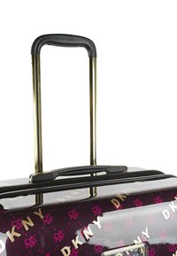 Maleta rígida de color púrpura oscuro con patrón "DKNY" en rosa y dorado. Cuenta con un asa retráctil dorada y un asa superior para llevar.