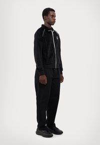 Veste zippée en velours noir avec garniture blanche, associée à un pantalon noir et des chaussures noires, mettant en avant une coupe décontractée et un design minimaliste.