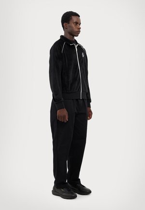 Black velour zip-up jacket with white piping, paired with black trousers and black shoes, showcasing a relaxed fit and minimal design.