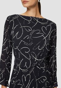 Schwarzes Kleid mit langen Ärmeln, das aus einem strukturierten Stoff besteht und herzförmige weiße Muster sowie handschriftliche Akzente im Design aufweist.