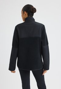 Zwarte fleecejas met een gladde nylon bovenpaneel, lange mouwen en een hoge kraag. De textured body vormt een contrast met de slanke stof.