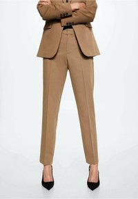 Une personne portant un costume beige avec un pantalon ajusté à la cheville et des escarpins noirs à bout pointu, se tenant bras croisés sur un fond blanc.