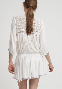 Vit klänning med trekvartsärmar, texturerad spets bakpanel, rynkad midja och blå kant längs nederkanten och ärmsluten.