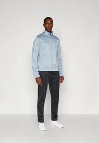 Gaiši zils rāvējslēdzēja jaka ar augstu apkakli un elastīgiem aprocēm, kopā ar melnām biksēm un baltām sporta kurpēm. Gluda, spīdīga auduma.