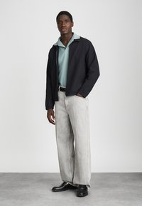 Čierna bunda s textúrovanou tkaninou, svetlomodrá polokošeľa, šedé široké nohavice a čierne topánky. Jednoduchý, moderný dizajn a farebný kontrast.