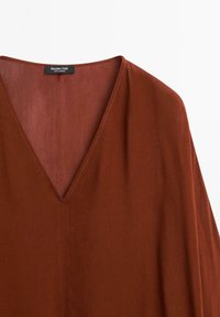 Blusa color ruggine con scollatura a V profonda, tessuto leggero e maniche corte. Presenta cuciture minime e un'etichetta di marca sul collo.