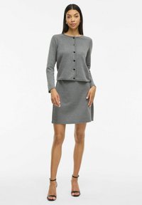 Ensemble en tricot gris composé d'un haut à boutons à manches longues et d'une jupe droite, présentant un tissu texturé et une silhouette décontractée.