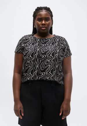 Frau mit geflochtenem Haar, die goldene Creolen, ein kurzärmeliges Top mit abstraktem schwarz-weißem Muster und schwarze Shorts trägt, stehend in neutraler Pose.