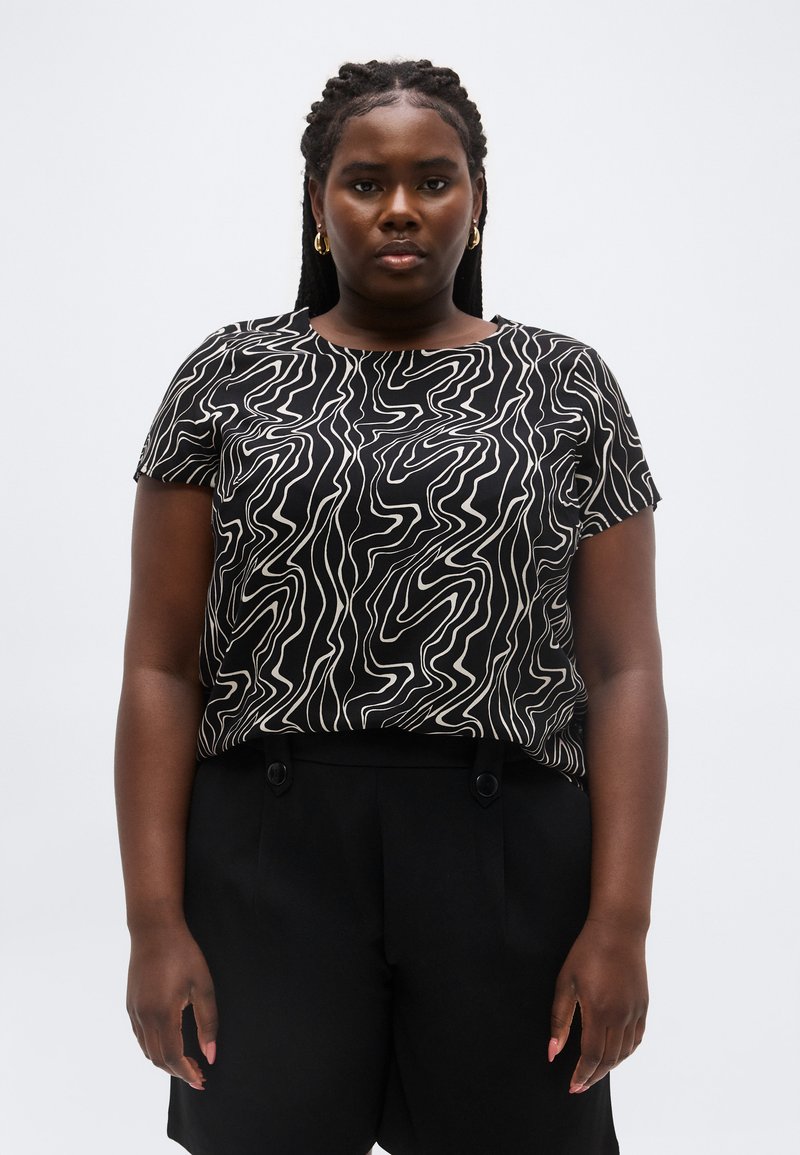 Γυναίκα με κοτσίδες στα μαλλιά, φορά χρυσούς κρίκους στα αυτιά, μπλούζα με κοντά μανίκια σε ασπρόμαυρο αφηρημένο σχέδιο και μαύρο σορτσάκι, στέκεται ουδέτερα.