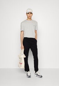 Gray short-sleeve T-shirt with small logo, black pants, white cap with text, cream tote bag with graphics, and gray and black sneakers.