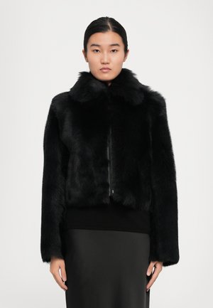 Vrouw in een zwarte bontjas met hoge kraag, gecombineerd met een zwart top en rok, staande tegen een effen lichte achtergrond.