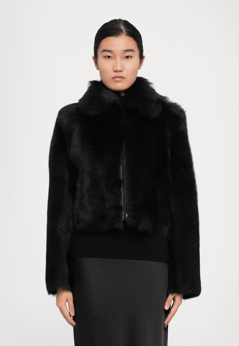 Woman wearing a black fur jacket with a high collar, paired with a black top and skirt, standing against a plain light background.