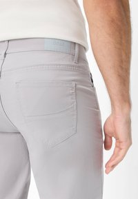 Pantaloni grigi chiari con una tasca posteriore e un sottile dettaglio del logo, texture liscia e design aderente. Tessuto morbido con cuciture pulite.