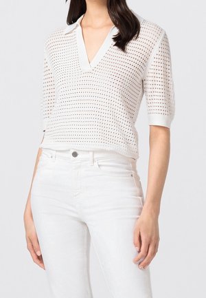 Femme portant un haut en maille blanc à manches courtes perforé avec un col en V, associé à un jean blanc taille haute, sur un fond clair.