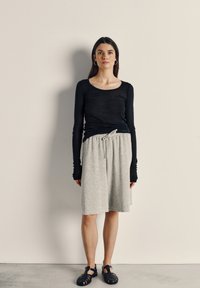 Žena s dugom tamnom kosom, odjevena u crni dugih rukava, svijetlosivu suknju dužine do koljena s vezicom i crne sandale zatvorenih prstiju, stoji nasuprot bež zidu.