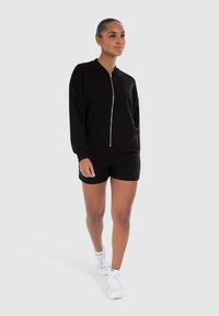 Veste bomber noire zippée associée à un short noir, tous deux en tissu doux et lisse. Le modèle porte des chaussures de sport blanches. Design simple et épuré.