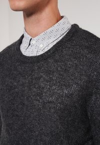 Pull en maille gris foncé avec un col rond, porté sur une chemise gris clair présentant un motif subtil de points noirs et un col boutonné.