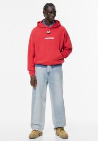 Persoon met gevlochten haar, gekleed in een rode "STWD STUDIO" hoodie, een gestreept shirt, lichtblauwe baggy jeans en beige laarzen op een witte achtergrond.