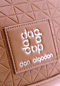 Cuero texturizado marrón con un patrón geométrico, presentando una etiqueta de logo cosida en blanco, "don algodon" debajo de un diseño estilizado.