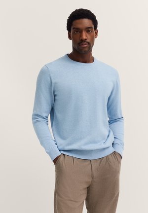 Mann mit Händen in den Taschen, der einen hellblauen Pullover und beidgefarbene Hosen vor einem einfarbigen hellen Hintergrund trägt.