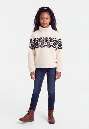 Fille aux cheveux bouclés portant un pull en polaire crème avec un motif floral noir, un jean skinny foncé et des bottes marron à lacets, debout sur fond blanc.