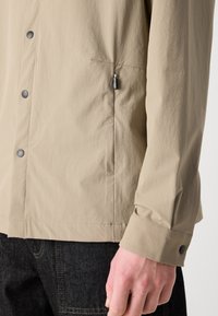Giacca leggera beige con tasca con zip, bottoni a pressione neri e tessuto testurizzato. I polsini sono rifiniti con chiusure a pressione.